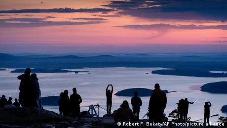 Amanecer en el Parque Nacional de Acadia, en Maine, EE. UU. El cielo está teñido de violeta y rosado, y las personas admiran la salida del sol junto al lago.