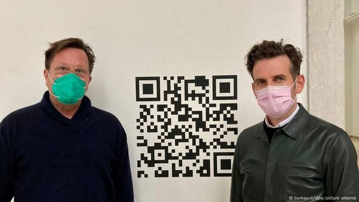 Arno Brandlhuber and Olaf Grawert wearing masks stand next to a giant QR code