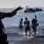 Soldado sinaliza para jovens que saem do mar, barcos ao fundo