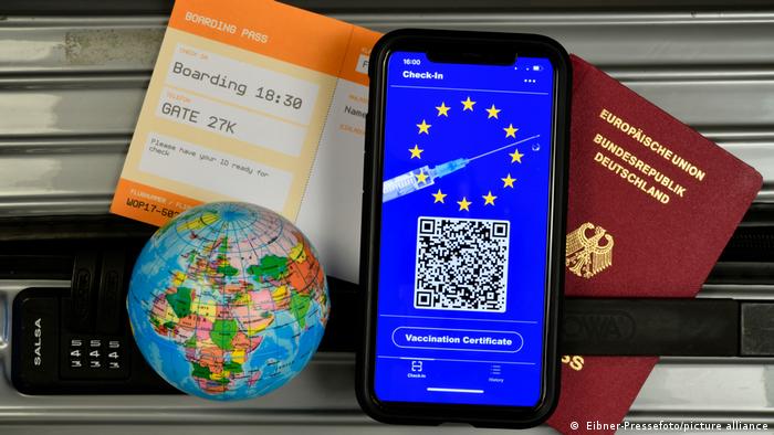 Ein Smartphone mit einer Impfpass-Darstellung, ein deutscher Reisepass, ein Flugticket und ein Mini-Globus liegen auf einem Koffer