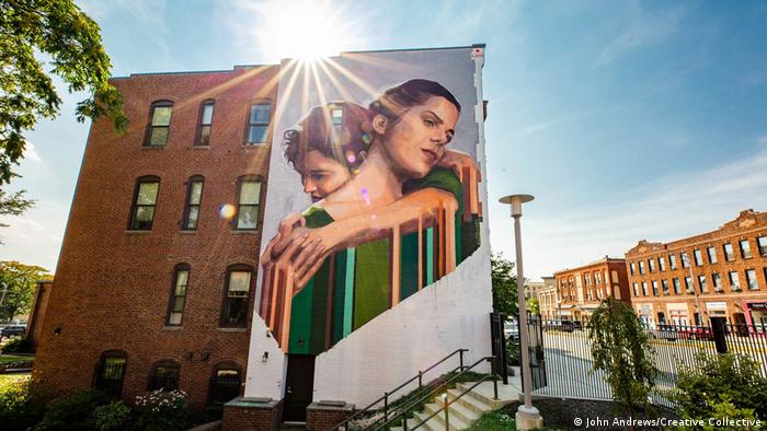 “Sororidad”, de Mariela Arjas: un mural sobre la fachada de un edificio. Una mujer abraza a otra, ambas vestidas en tonos de verde.