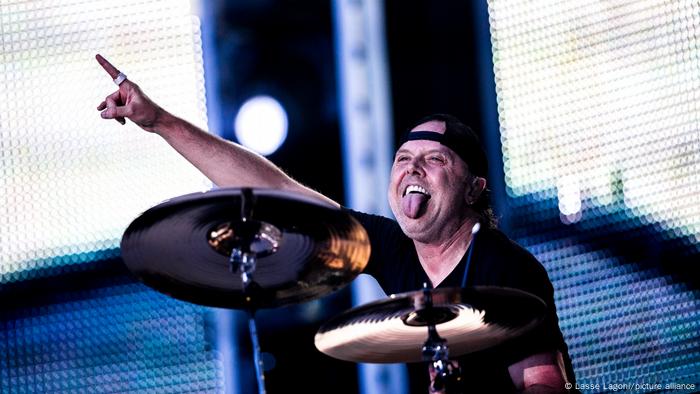 Lars Ulrich sticking his tongue out and pointing his hand while sitting behind drums. 