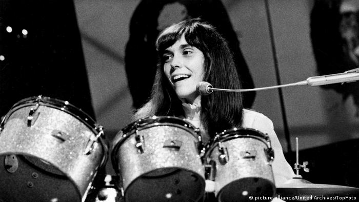 A black-and-white image of Karen Carpenter behind the drums, smiling. 