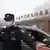 Um policial tenta tirar a visão do fotógrafo enquanto um carro chega ao Instituto de Virologia de Wuhan, na China.