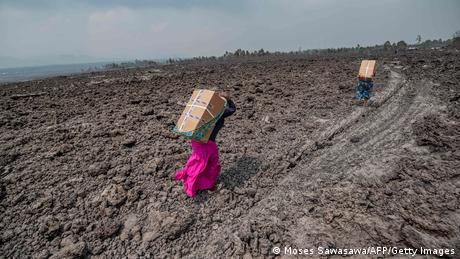 Deux femmes portent de grands cartons sur le dos et marchent sur un champ de lave refroidi
