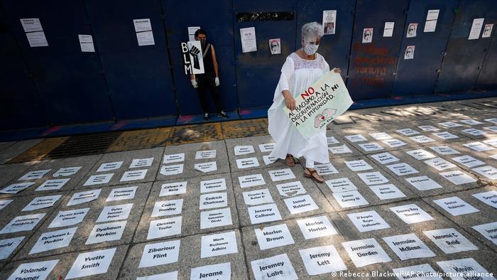 Eine Frau hält ein Schild aus Spanisch gegen Rassissmus, Diskrikminierung und Straffreiheit, am Boden zig Blätter mit Namen von Gewaltopfern