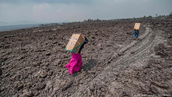 Residents wearing bright dresses carry goods on their backs, bent forward in field of ash