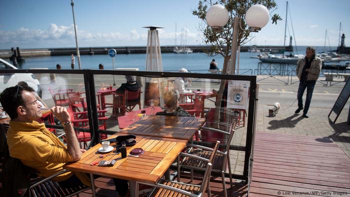 Homem sentado em uma mesa externa de um café. Sobre a mesa está uma xícara de café. Há uma barreira de vidro na frente dele. Ao fundo está o mar. 