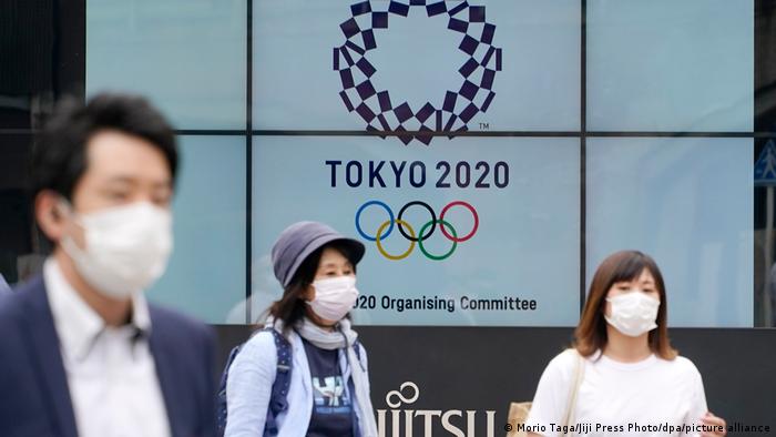 Japanac i dvije Japanke stoje ispred video screena sa logom Olimpijskih igara u Tokiju
