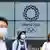 Japanac i dvije Japanke stoje ispred video screena sa logom Olimpijskih igara u Tokiju