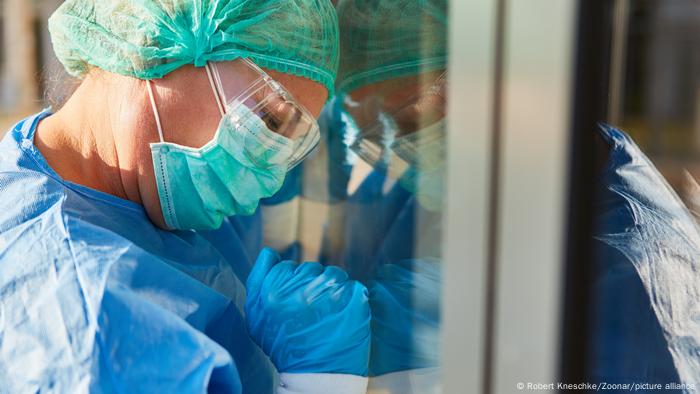 Mediziner mit Schutzkleidung bei Coronavirus Pandemie lehnt ermüdet Kopf an Fenster