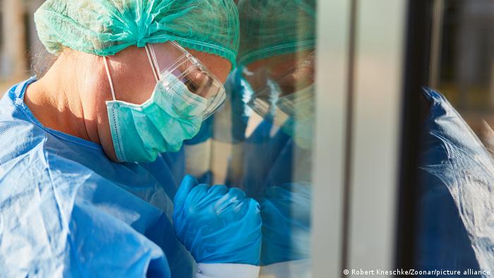 Mediziner mit Schutzkleidung bei Coronavirus Pandemie lehnt ermüdet Kopf an Fenster