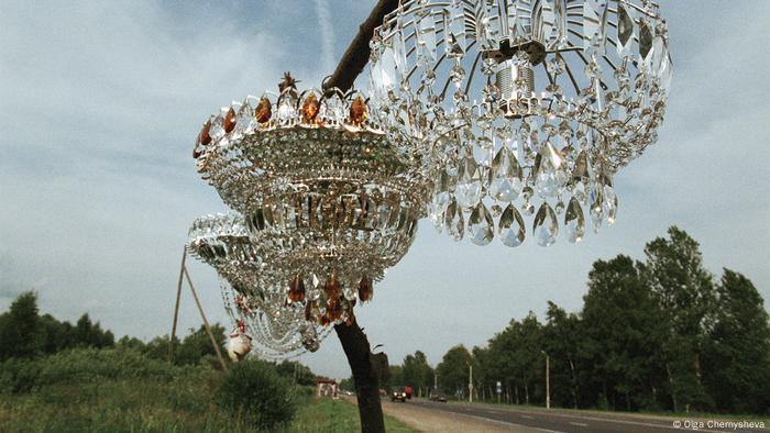 Olga Chernysheva artwork 'On the Sidelines': A photo of three chandeliers hanging from a roadside tree.