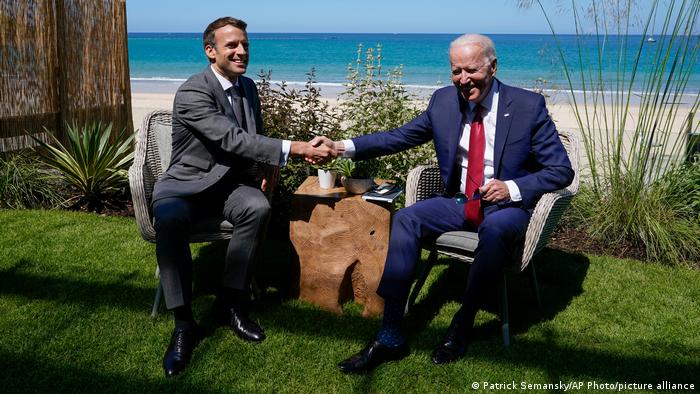 Joe Biden and Macron shake hands while posing for a photograph with the sea and sandy beach behind them