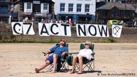 Ein älteres, weißes Paar sonnt sich bequem am Strand in Liegestühlen, im Hintergrund ein Plakat G7 ACT NOW