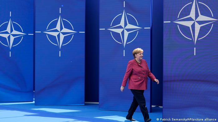 Kanzlerin Merkel läuft beim NATO-Gipfel an einer Wand mit vier großen NATO-Flaggen entlang
