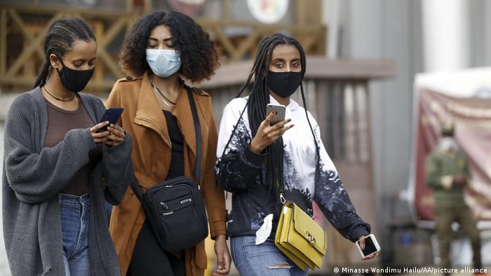 Drei Frauen mit Maske laufen über eine Straße und tippen auf ihren Smartphones