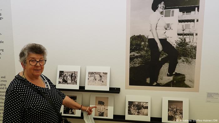 Asimina Paradissa steht - mit Brille und kurzen grauen Haaren - vor einer Wand mit Fotos und deutet auf ein Bild, das sie als junge Frau zeigt. Asimina Paradissa steht - mit Brille und kurzen grauen Haaren - vor einer Wand mit Fotos und deutet auf ein Bild, das sie als junge Frau zeigt.