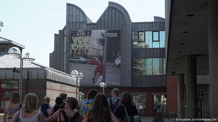 Eine Gruppe von Schülern geht auf das Museum Ludwig zu. An der Fassade hängt ein Plakat zur Ausstellung: „Vor Ort: Fotogeschichten zur Migration“. Eine Gruppe von Schülern geht auf das Museum Ludwig zu. An der Fassade hängt ein Plakat zur Ausstellung: „Vor Ort: Fotogeschichten zur Migration“.