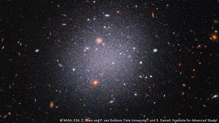 Esta instantánea del telescopio espacial Hubble revela una inusual galaxia transparente. La gigantesca bola de algodón cósmica es tan difusa y sus antiguas estrellas están tan dispersas que las galaxias lejanas del fondo pueden verse a través de ella. Denominada galaxia ultradifusa, este bicho raro galáctico es casi tan ancho como la Vía Láctea, pero contiene sólo una 1/200 parte del número de estrellas que nuestra galaxia.