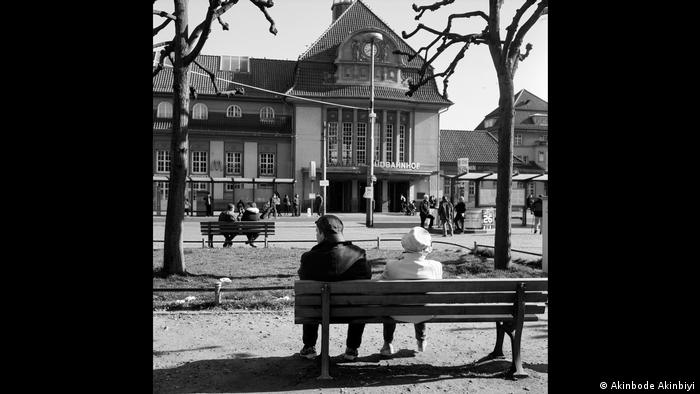 A couple sits on a bench in front of a railway station.