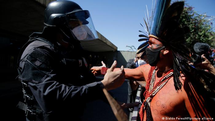 An Indigenous leader wearing a feathered head dress faces off with riot police