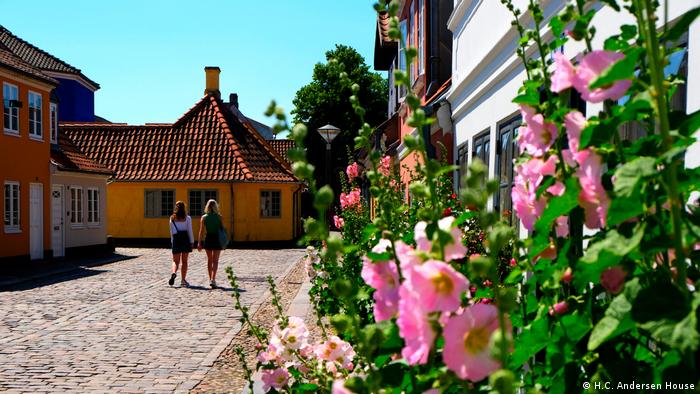 Two women walk down a cobbled street, buildings to the left and right, flowers