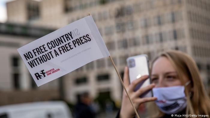 A woman wearing a mask holds a cellphone and a small flag showing the slogan No free country without a free press and the Reporters Without Borders logo