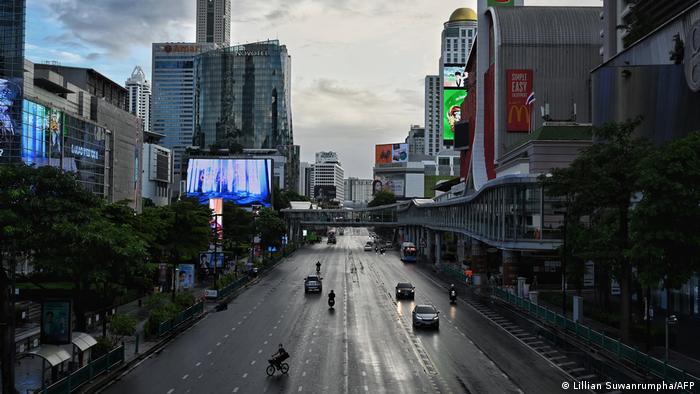 A few cars and bikes travel along a six-lane road in the middle of Bangkok. There are big buildings on either side and a bridge over the road. There is lots of empty space on the road.