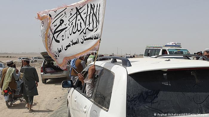 people in a white car wave the white Taliban flag as they drive through a dusty and busy border crossing