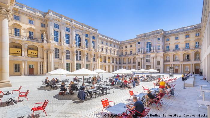 People sitting at cafe tables with umbrellas in the inner courtyard of the Humboldt Forum.