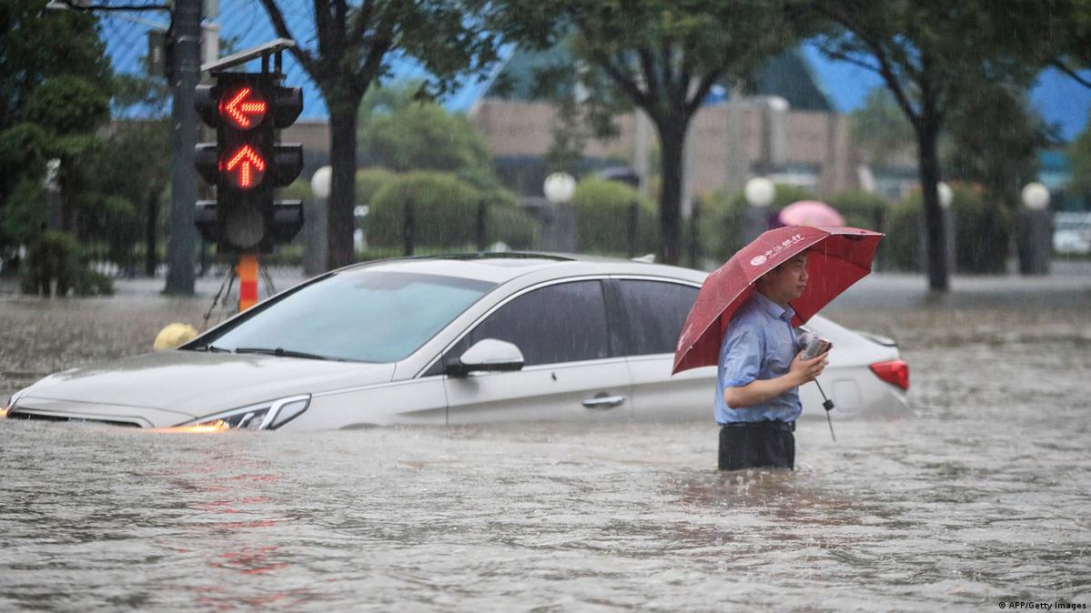 Ein Mann mit Regenschirm steht bis zur Hüfte in den Fluten. Im Hintergrund ein Auto, das in den Fluten feststeckt