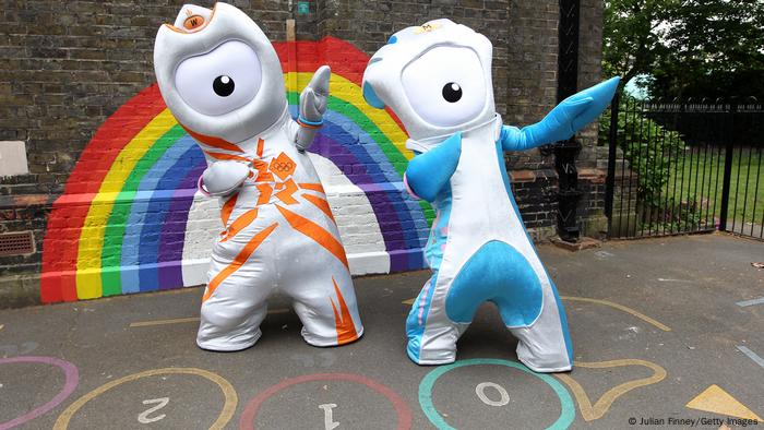 The two Olympic mascots Wenlock (left) and Mandeville (right) stand in front of a brick wall painted with a rainbow. 