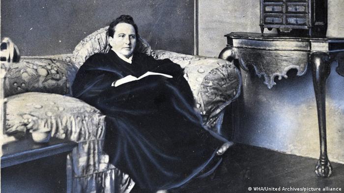 Author and poet, Gertrude Stein dressed in dark clothes, sitting on a sofa with a side table to the right of the picture