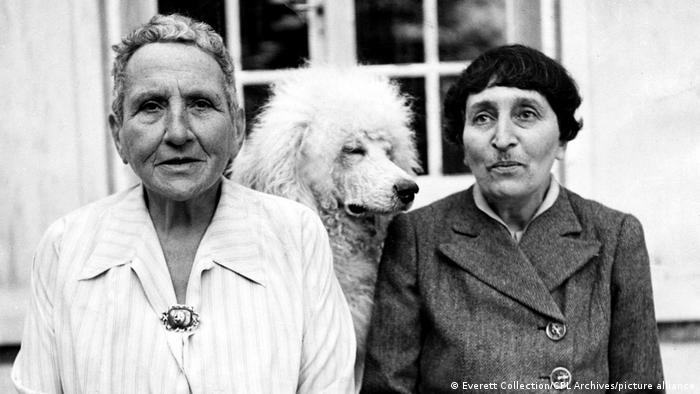 Author Gertrude Stein dressed in a striped top with a brooch on, and Alice B. Toklas her partner dressed in a dark single breates coat. Their furry white dog, Basket, sits between them