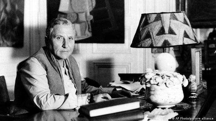 Author Gertrude Stein, looks into the camera while seated at her desk that has books, a pot of flowers and geomatric-patterned lampshade
