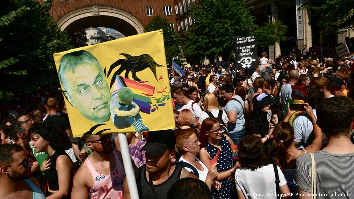 People taking part in a gay pride parade in Budapest with one holding a sign depicting Hungarian Prime Minister Viktor Orban with a rainbow flag and a sad, isolated child