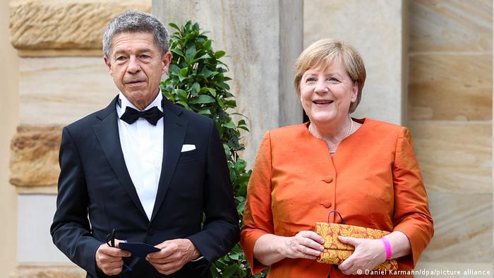 Angela Merkel in an orange blazer stands next to her husband Joachim Sauer in a tuxedo