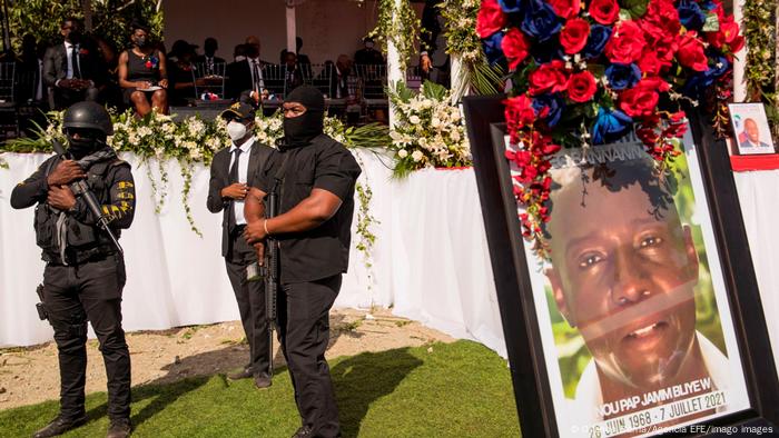 Two armed and masked guards stand close to a photo of Jovenel Moise at his funeral