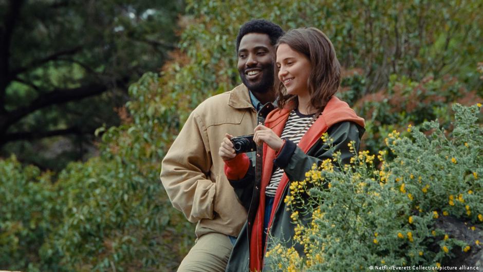 Film still | BECKETT | John David Washington and Alicia Vikander, a man and a woman stand among shrubs