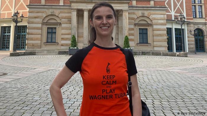 A woman stands in front of an elegant building entryway wearing a shirt that says 'Keep calm and play Wagner-Tuba'