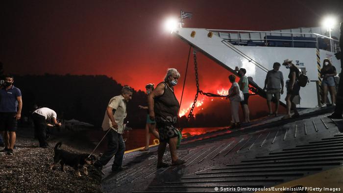 Passageiros sobem em embarcação, com fogo florestal ao fundo