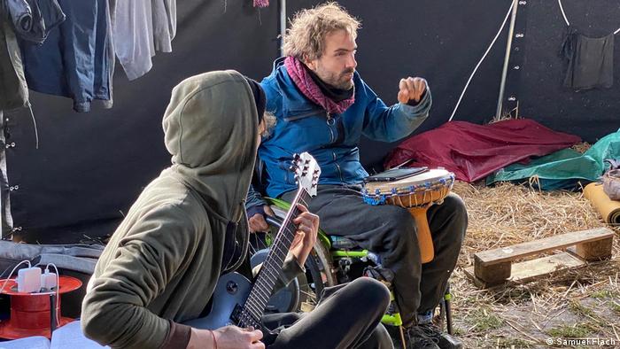 Umweltaktivist und Rollstuhlfahrer Samuel Flach mit einer Trommel zwischen den Knien neben einem Gitarrenspieler in einem Zelt im Dannenröder Forst