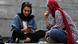 Two women in Iran are sitting on steps, looking at a smartphone.