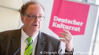 Olaf Zimmermann, a man in a grey suit with a green tie in front of a panel that reads 'Deutscher Kulturrat' (German Cultural Council)