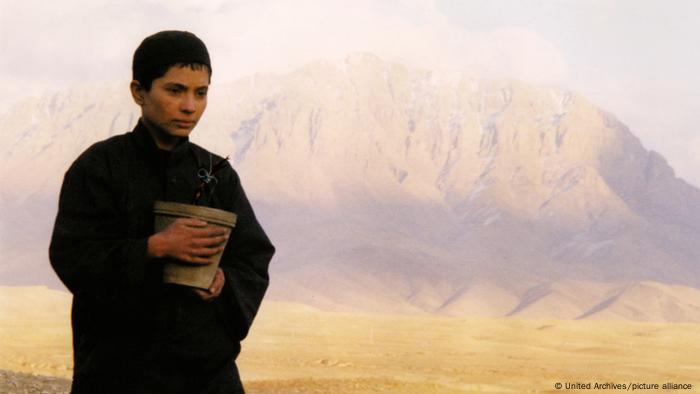 Szene aus dem Film Osama, mit einem jungen Mann, der eine Blumenvase hält, mit einer Bergkette im Hintergrund.