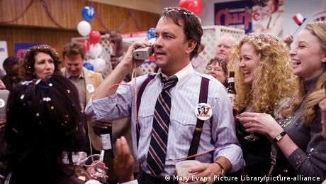 Film still from 'Charlie Wilson's War' - Tom Hanks speaking on an 1980s cell phone at an election celebration, various people smiling in the background.