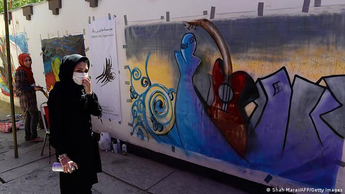 Shamsia Hassani steht mit einer Sprühdose in der Hand vor einem Wandgemälde