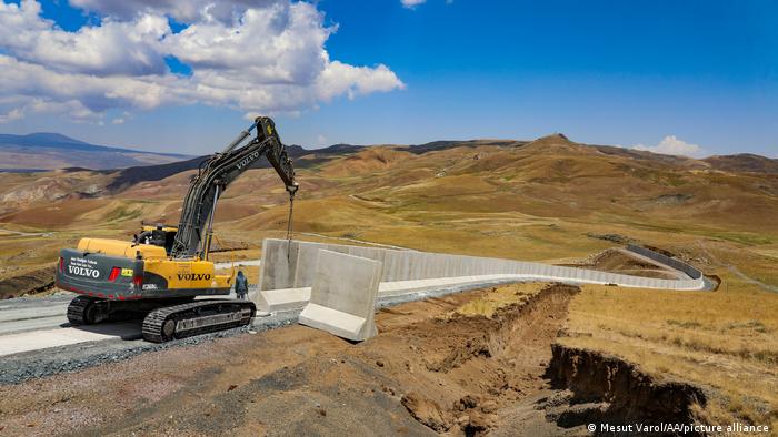 Ein Raupenkran platziert Beton-Fertigteile als Mauer in karger und unbewohnter Landschaft an der türkisch-iranischen Grenze