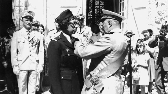 A military man pins an award on the jacket of a woman who is also wearing a military uniform